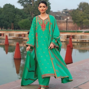 May include: A teal green embroidered ensemble featuring a long-sleeved tunic, matching pants, and a shawl. The outfit is adorned with red and gold floral embroidery. The model is wearing red shoes. The background includes a body of water and a building.