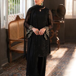 May include: A black ensemble featuring a long-sleeved tunic with intricate embroidery on the sleeves and a matching scarf. The outfit includes wide-leg pants and is accessorized with jewelry. The setting is a room with a patterned tile floor and a vintage chair.