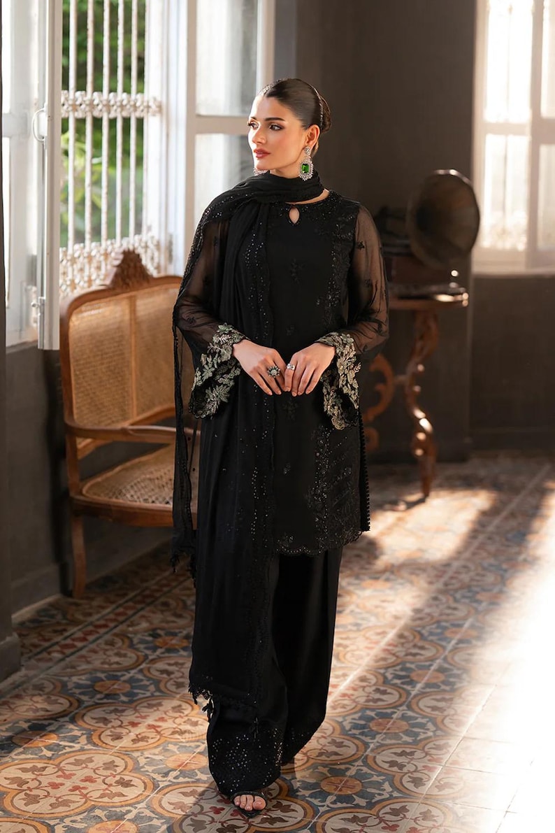 May include: A black ensemble featuring a long-sleeved tunic with intricate embroidery on the sleeves and a matching scarf. The outfit includes wide-leg pants and is accessorized with jewelry. The setting is a room with a patterned tile floor and a vintage chair.