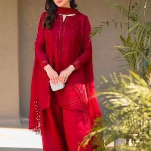 May include: A woman wearing a red embroidered dress with a matching dupatta and pants. The dress has a high neckline and long sleeves. The dupatta is draped over her shoulders and has a lace trim. The pants are wide-legged and flowy.