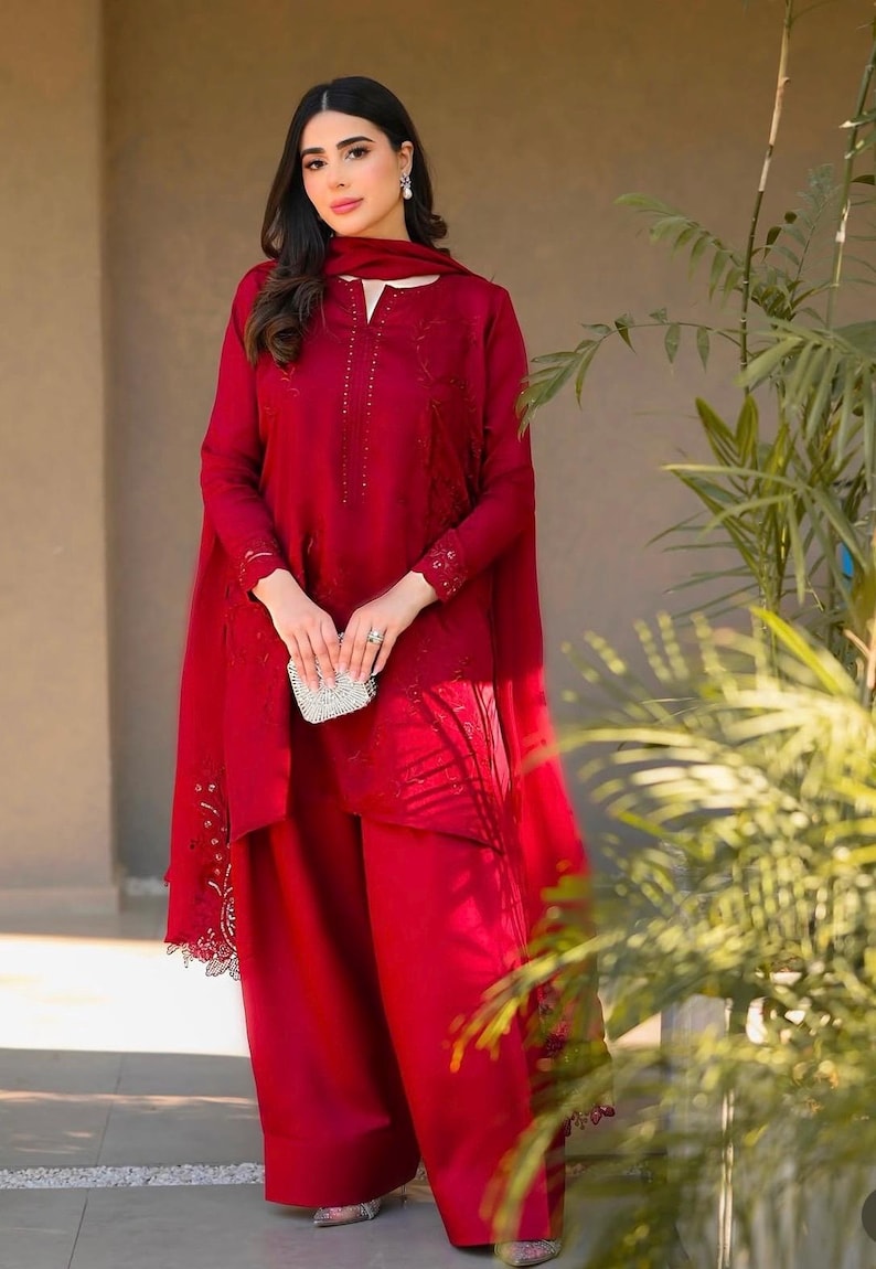 May include: A woman wearing a red embroidered dress with a matching dupatta and pants. The dress has a high neckline and long sleeves. The dupatta is draped over her shoulders and has a lace trim. The pants are wide-legged and flowy.