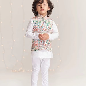 May include: A young boy wearing a white kurta and a colorful patterned waistcoat. He is standing on a white background with string lights in the background.
