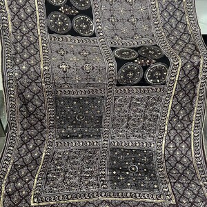 K&ouml;nnte beinhalten: Ein dunkelviolettes und schwarzes Textil mit komplizierten Mustern. Das Design zeigt geometrische Formen, florale Motive und kreisf&ouml;rmige Elemente, die alle in einer helleren Farbe umrandet sind. Der Rand hat ein detailliertes, sich wiederholendes Muster.