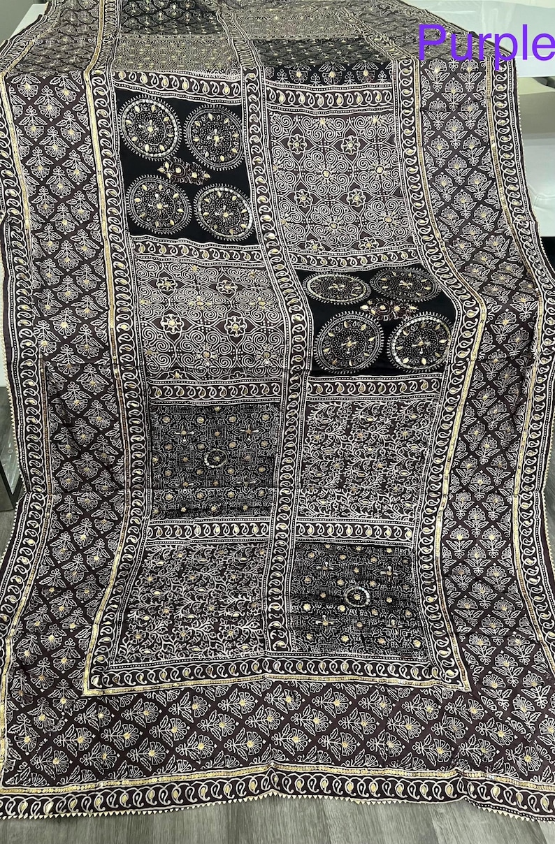 K&ouml;nnte beinhalten: Ein dunkelviolettes und schwarzes Textil mit komplizierten Mustern. Das Design zeigt geometrische Formen, florale Motive und kreisf&ouml;rmige Elemente, die alle in einer helleren Farbe umrandet sind. Der Rand hat ein detailliertes, sich wiederholendes Muster.