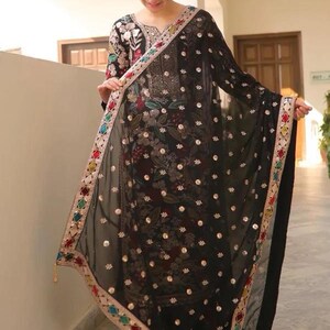 May include: A black embroidered salwar kameez with a matching dupatta. The outfit features floral and sequin embellishments. The dupatta has a colorful border with geometric patterns and tassels. The outfit is worn by a person.