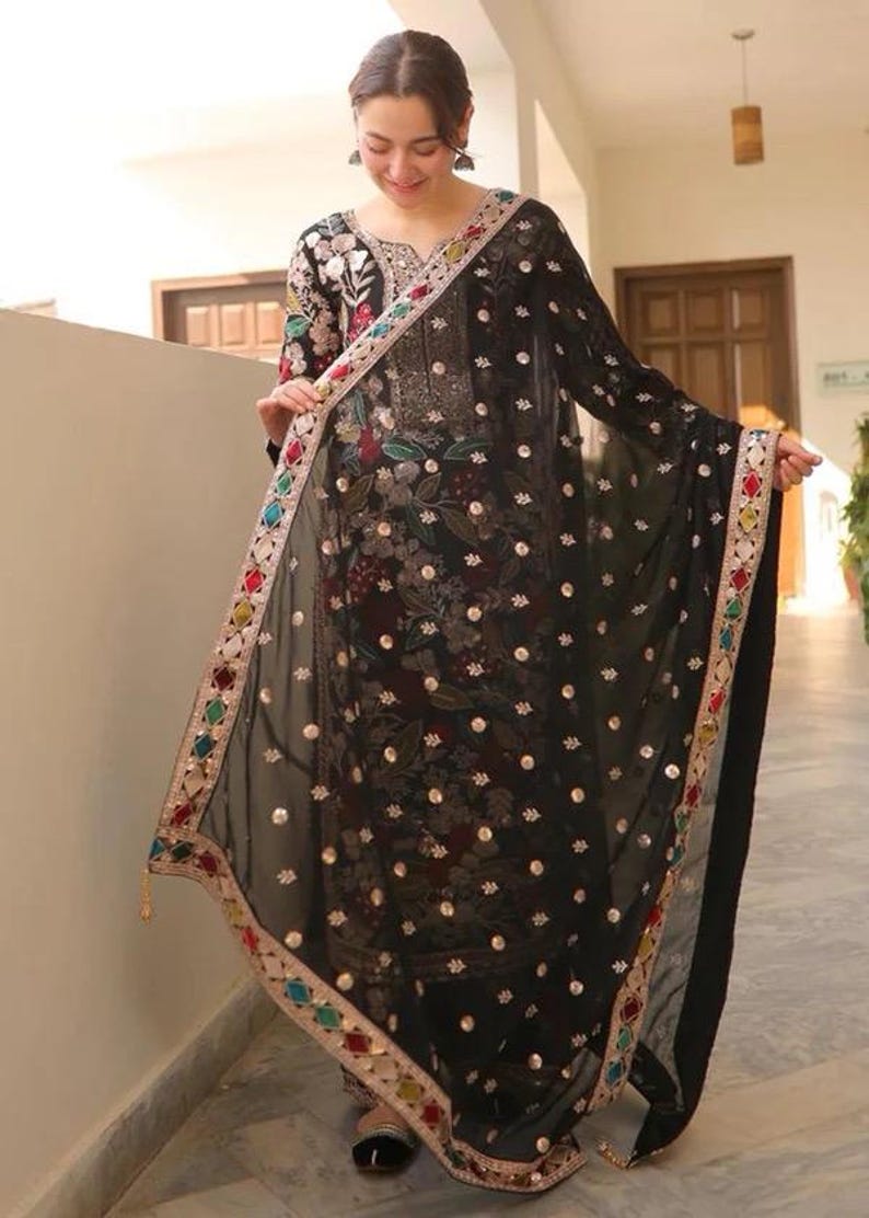 May include: A black embroidered salwar kameez with a matching dupatta. The outfit features floral and sequin embellishments. The dupatta has a colorful border with geometric patterns and tassels. The outfit is worn by a person.