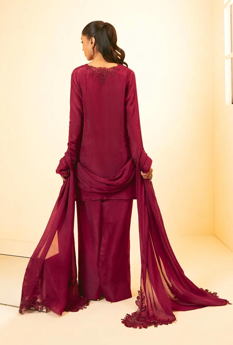 May include: A burgundy-colored outfit with a long-sleeved top and wide-legged pants. The top has a delicate lace trim around the neckline. The outfit is paired with a matching sheer dupatta with lace trim.
