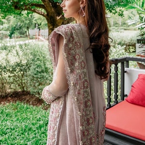 May include: A woman wearing a light pink and gray saree with intricate embroidery and pearl accents. The saree has a gradient effect, transitioning from a darker pink at the bottom to a lighter gray at the top. The woman is standing in front of a lush green garden.