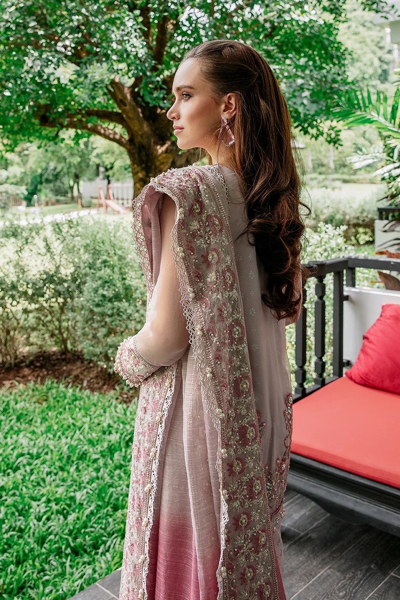 May include: A woman wearing a light pink and gray saree with intricate embroidery and pearl accents. The saree has a gradient effect, transitioning from a darker pink at the bottom to a lighter gray at the top. The woman is standing in front of a lush green garden.