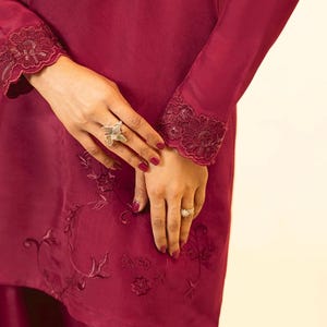 May include: A maroon long-sleeved top with lace trim at the cuffs and embroidered floral designs. The top is paired with a matching maroon skirt.