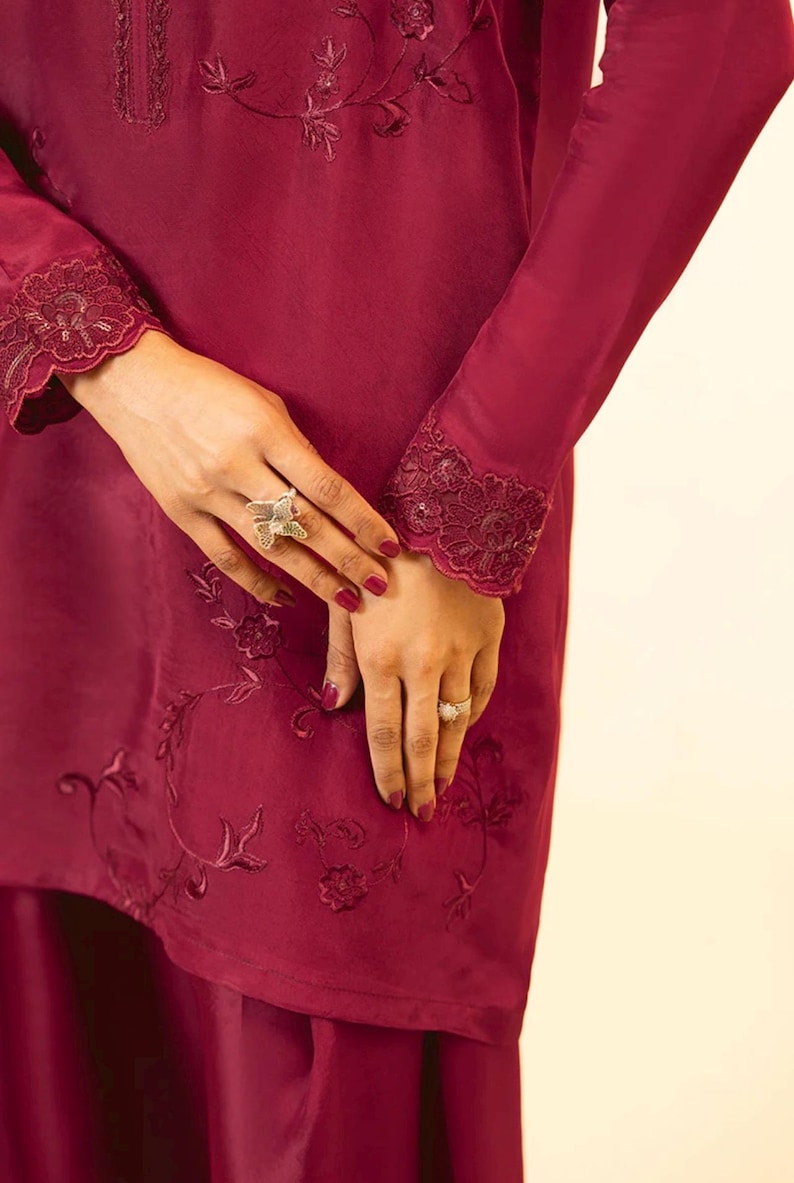 May include: A maroon long-sleeved top with lace trim at the cuffs and embroidered floral designs. The top is paired with a matching maroon skirt.