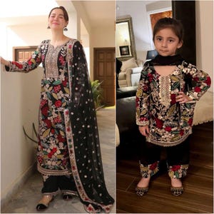 May include: Matching black embroidered outfits. The adult outfit includes a long-sleeved tunic, matching pants, and a sheer black scarf. The tunic and pants are adorned with floral embroidery in red, gold, and green. The child's outfit mirrors the adult's design.