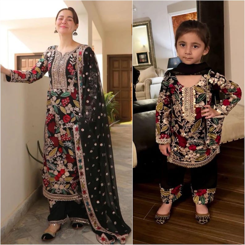 May include: Matching black embroidered outfits. The adult outfit includes a long-sleeved tunic, matching pants, and a sheer black scarf. The tunic and pants are adorned with floral embroidery in red, gold, and green. The child's outfit mirrors the adult's design.