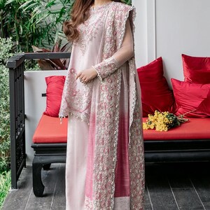 May include: A woman wearing a pink and white embroidered dress with a matching dupatta. The dress has a long, flowing skirt and a fitted bodice. The dupatta is draped over the woman's shoulders and is decorated with intricate embroidery. The woman is standing in front of a red couch with pillows.