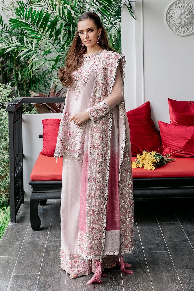 May include: A woman wearing a pink and white embroidered dress with a matching dupatta. The dress has a long, flowing skirt and a fitted bodice. The dupatta is draped over the woman's shoulders and is decorated with intricate embroidery. The woman is standing in front of a red couch with pillows.