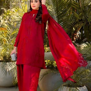 May include: A woman wearing a red embroidered outfit with a matching dupatta. The outfit features intricate floral designs and embellishments. The woman is standing in front of a lush green background.