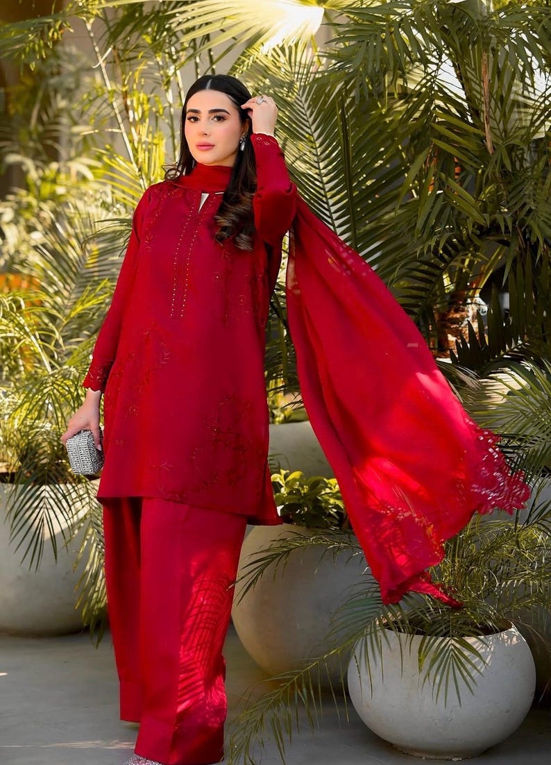May include: A woman wearing a red embroidered outfit with a matching dupatta. The outfit features intricate floral designs and embellishments. The woman is standing in front of a lush green background.