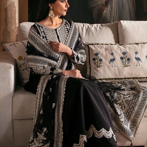 May include: A woman wearing a black embroidered dress with silver embellishments and a matching shawl. She is sitting on a white couch with a floral patterned pillow.