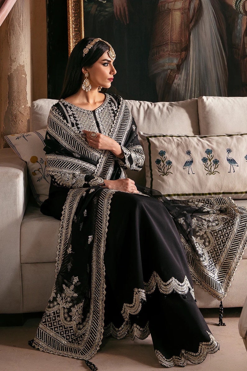 May include: A woman wearing a black embroidered dress with silver embellishments and a matching shawl. She is sitting on a white couch with a floral patterned pillow.