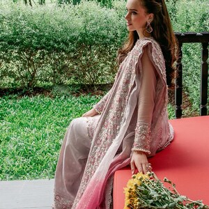 May include: A woman wearing a long, flowing, pink and gray embroidered dress with a matching shawl. She is sitting on a red bench with a bouquet of yellow flowers in her hand.