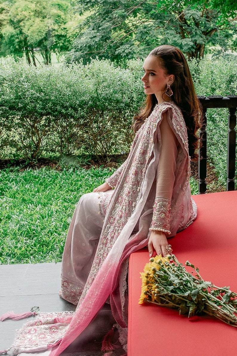 May include: A woman wearing a long, flowing, pink and gray embroidered dress with a matching shawl. She is sitting on a red bench with a bouquet of yellow flowers in her hand.