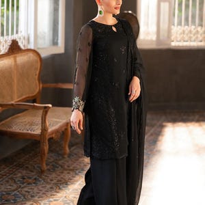 May include: Black formal outfit featuring a long-sleeved top with intricate embroidery and matching wide-leg pants. The ensemble includes a sheer shawl. The model is wearing a red flower in their hair and green earrings. The outfit is suitable for special occasions.