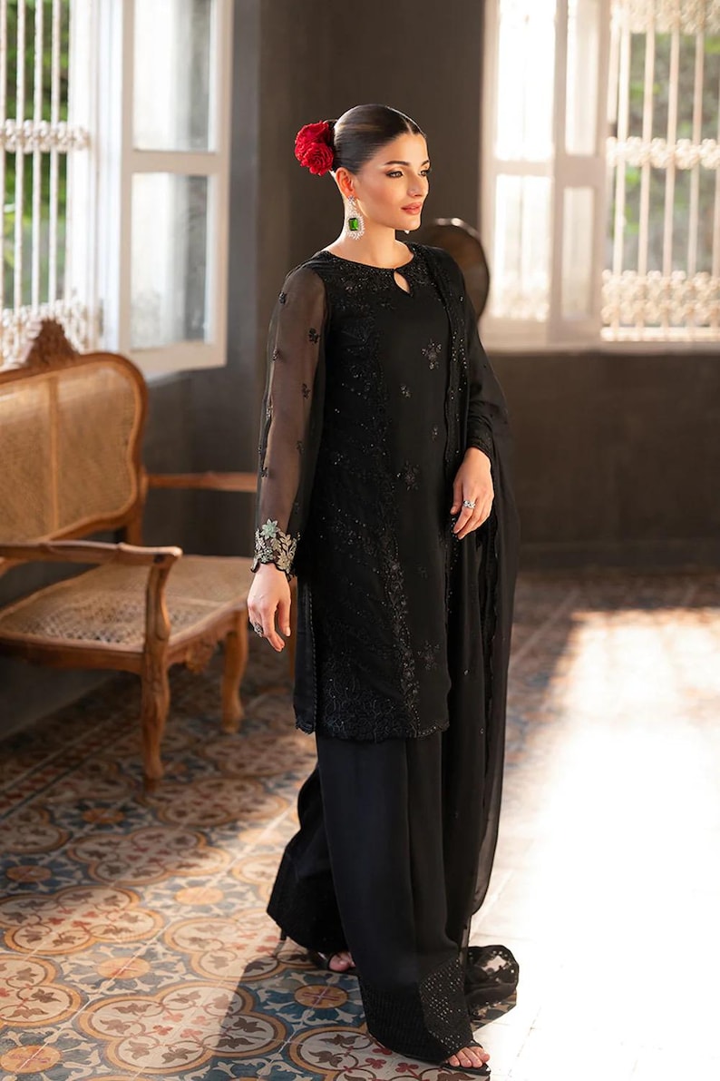 May include: Black formal outfit featuring a long-sleeved top with intricate embroidery and matching wide-leg pants. The ensemble includes a sheer shawl. The model is wearing a red flower in their hair and green earrings. The outfit is suitable for special occasions.