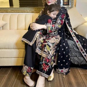 May include: A woman and a child are embracing while seated on a beige sofa. The woman wears a black outfit with floral embroidery and gold shoes. The child wears a black outfit. A black shawl with embroidery is draped over the woman's shoulder.