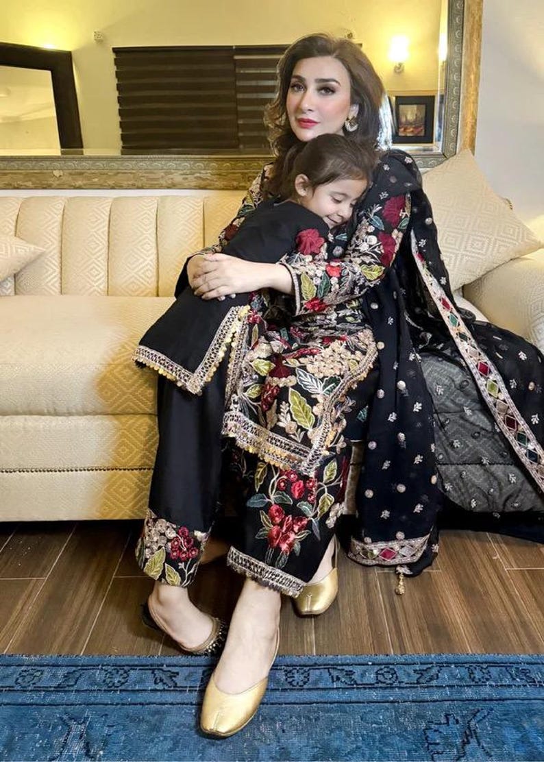 May include: A woman and a child are embracing while seated on a beige sofa. The woman wears a black outfit with floral embroidery and gold shoes. The child wears a black outfit. A black shawl with embroidery is draped over the woman's shoulder.