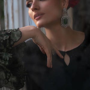 May include: A woman wearing a black dress with a keyhole neckline and floral embroidered sleeves. She is wearing large, square emerald and diamond earrings. Her hair is pulled back, and she has a red flower in her hair.