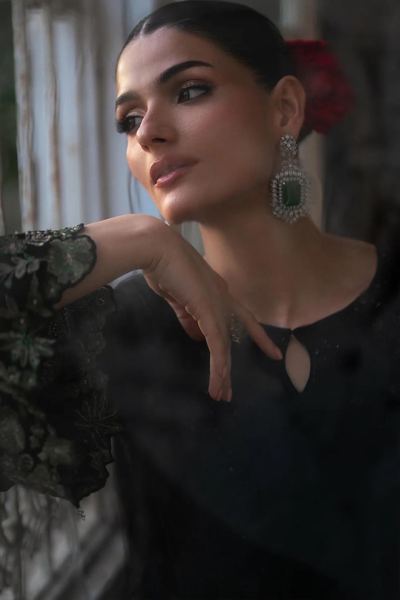 May include: A woman wearing a black dress with a keyhole neckline and floral embroidered sleeves. She is wearing large, square emerald and diamond earrings. Her hair is pulled back, and she has a red flower in her hair.