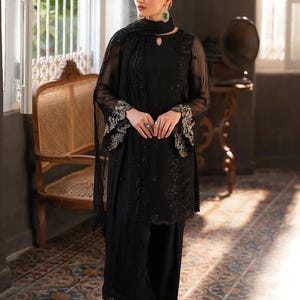 May include: A black ensemble featuring a long-sleeved tunic, wide-leg pants, and a matching scarf. The tunic and pants are adorned with intricate embroidery. The outfit is accessorized with jewelry. The model is standing in a room with a patterned tile floor.