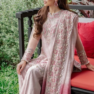 May include: A woman wearing a pink and red embroidered outfit with a long, flowing dupatta. She is sitting on a bench with a red cushion.