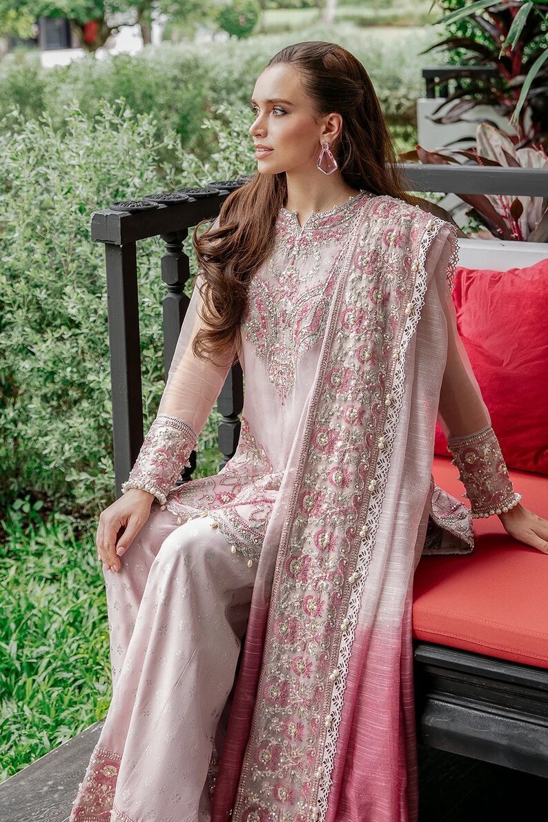 May include: A woman wearing a pink and red embroidered outfit with a long, flowing dupatta. She is sitting on a bench with a red cushion.