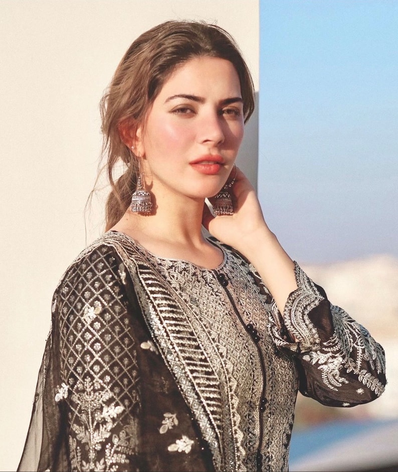 May include: A woman with long brown hair wearing a black and silver embroidered dress with a silver pendant earring on each ear.