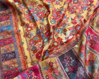 May include: A golden-yellow textile with a floral pattern in shades of red, pink, and blue. The fabric features a detailed border with rectangular panels and intricate designs. The textile is draped, highlighting its texture and colour variations.