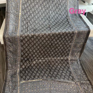 K&ouml;nnte beinhalten: Grauer Stoff mit sich wiederholendem wei&szlig;en Blumenmuster. Der Rand weist geometrische Muster und kleine reflektierende Akzente auf. Der Stoff ist &uuml;ber eine wei&szlig;e Oberfl&auml;che drapiert.