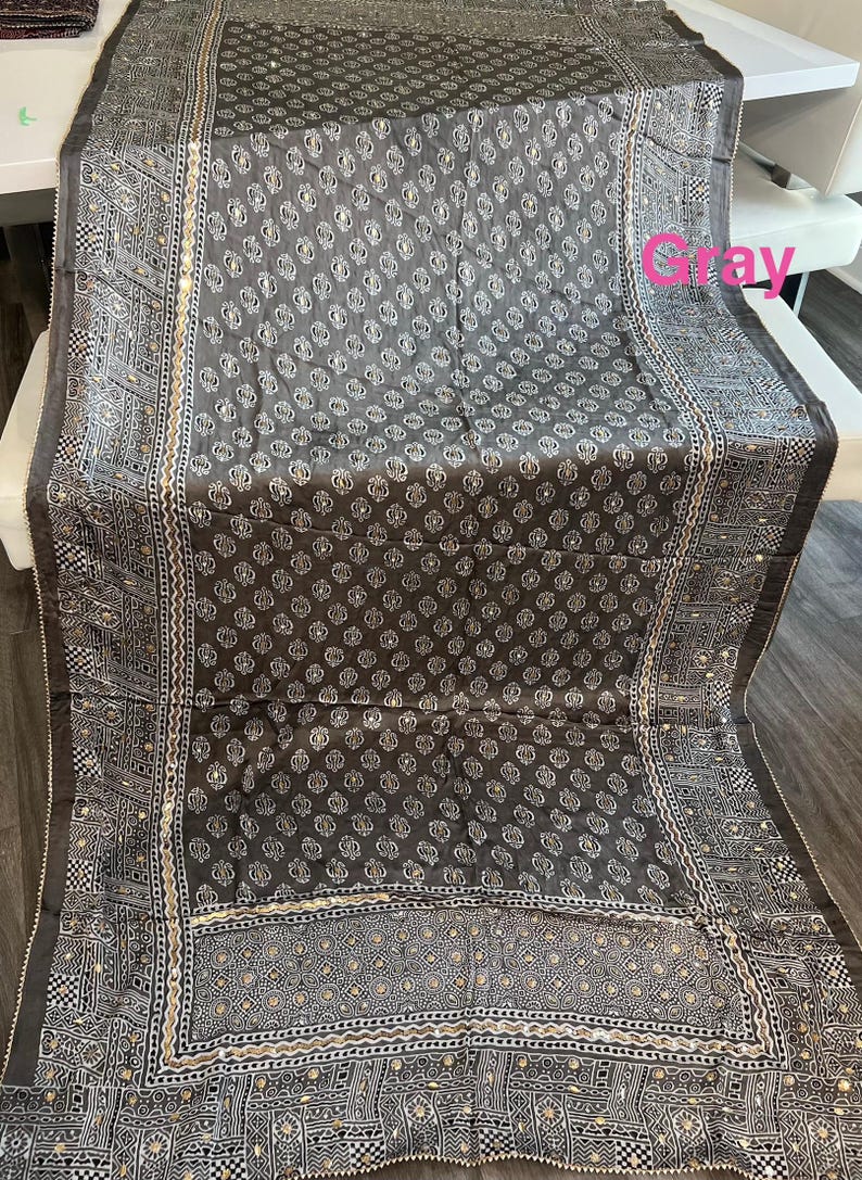 K&ouml;nnte beinhalten: Grauer Stoff mit sich wiederholendem wei&szlig;en Blumenmuster. Der Rand weist geometrische Muster und kleine reflektierende Akzente auf. Der Stoff ist &uuml;ber eine wei&szlig;e Oberfl&auml;che drapiert.