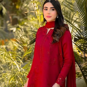 May include: A woman wearing a long-sleeved, red dress with intricate embroidery and a matching red scarf. The dress has a high neckline and a keyhole opening. The woman is standing in front of a green leafy background.