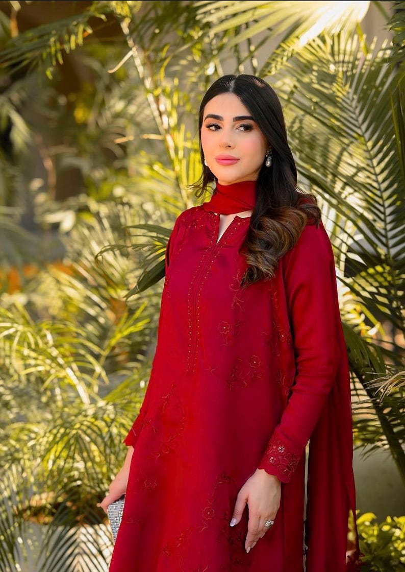 May include: A woman wearing a long-sleeved, red dress with intricate embroidery and a matching red scarf. The dress has a high neckline and a keyhole opening. The woman is standing in front of a green leafy background.