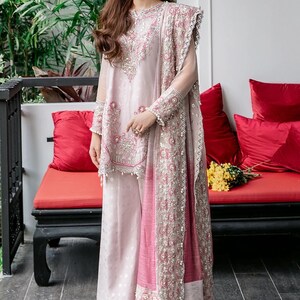 May include: A woman wearing a pink and white embroidered dress with a matching dupatta. The dress has a long, flowing skirt and a fitted bodice. The dupatta is decorated with intricate embroidery and has a tassel at the end.