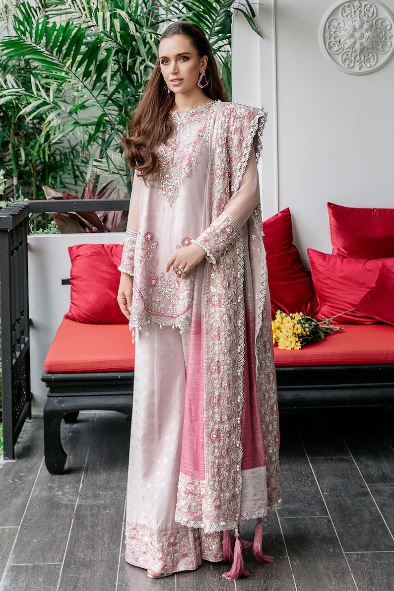 May include: A woman wearing a pink and white embroidered dress with a matching dupatta. The dress has a long, flowing skirt and a fitted bodice. The dupatta is decorated with intricate embroidery and has a tassel at the end.