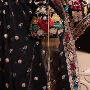 May include: Black fabric with gold floral accents and a matching pouch. The pouch features red and green floral embroidery. The garment has a decorative border with colorful diamond-shaped patterns and gold trim. A gold beaded chain hangs from the pouch.