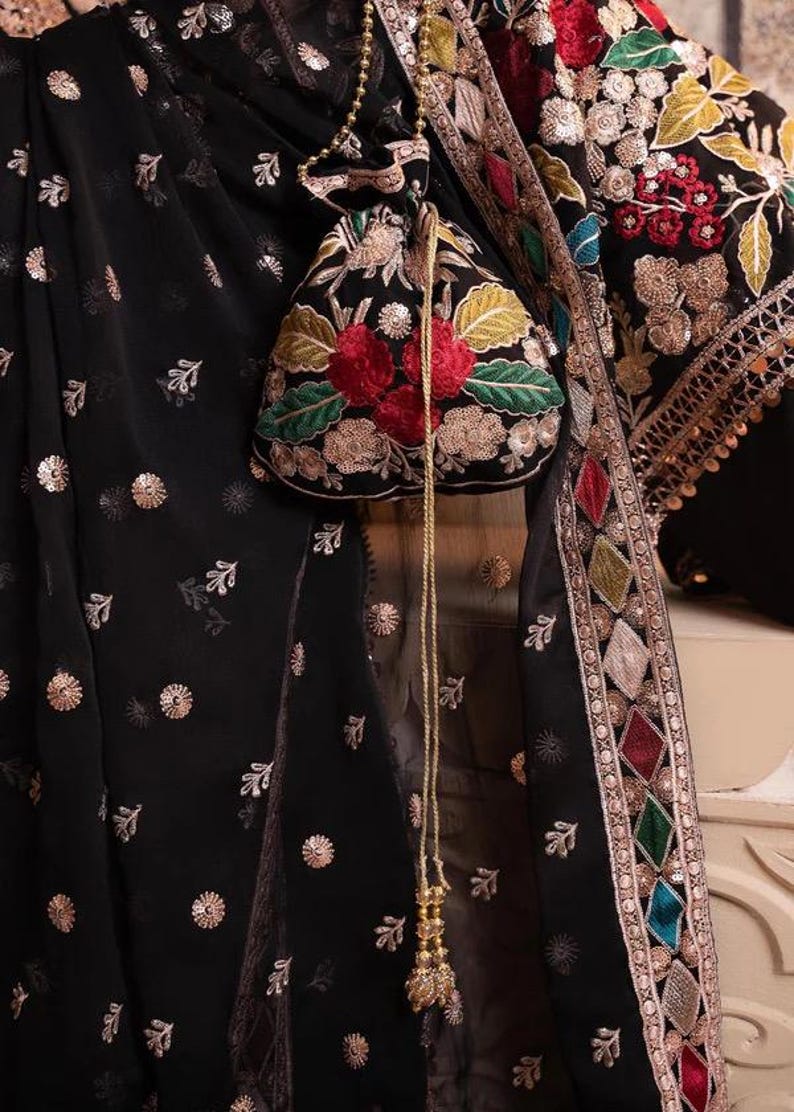 May include: Black fabric with gold floral accents and a matching pouch. The pouch features red and green floral embroidery. The garment has a decorative border with colorful diamond-shaped patterns and gold trim. A gold beaded chain hangs from the pouch.