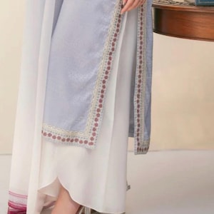 May include: A light blue and white ensemble featuring a long, flowing skirt and a matching top with intricate embroidered detailing. The outfit includes a sheer white overlay and is paired with clear heeled shoes.
