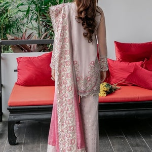 May include: A woman wearing a pink and white embroidered outfit with a matching pink dupatta. The outfit features intricate floral designs and pearl embellishments. The woman is standing in front of a red couch with pillows.