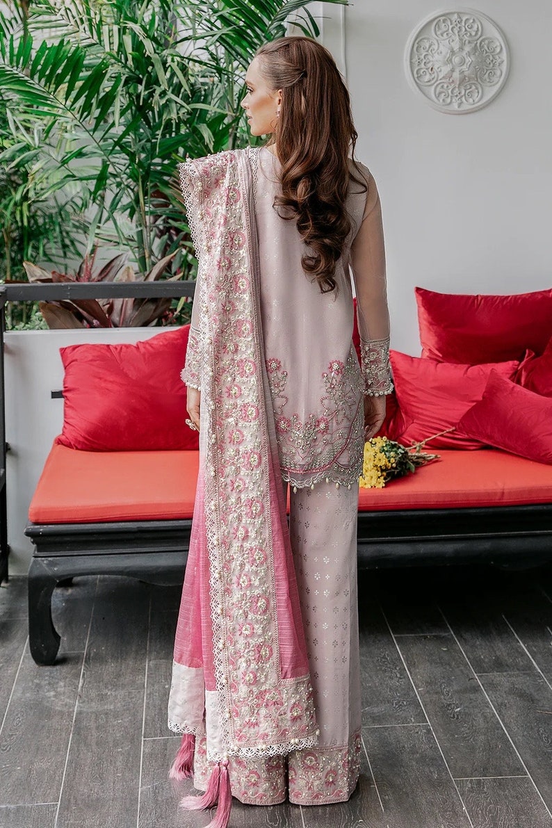 May include: A woman wearing a pink and white embroidered outfit with a matching pink dupatta. The outfit features intricate floral designs and pearl embellishments. The woman is standing in front of a red couch with pillows.