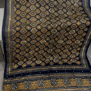 K&ouml;nnte beinhalten: Ein marineblaues Textil mit einem komplizierten Muster aus goldenen und wei&szlig;en floralen und geometrischen Formen. Das Textil hat einen dekorativen Rand und das Wort "Mustard" ist oben links zu sehen.