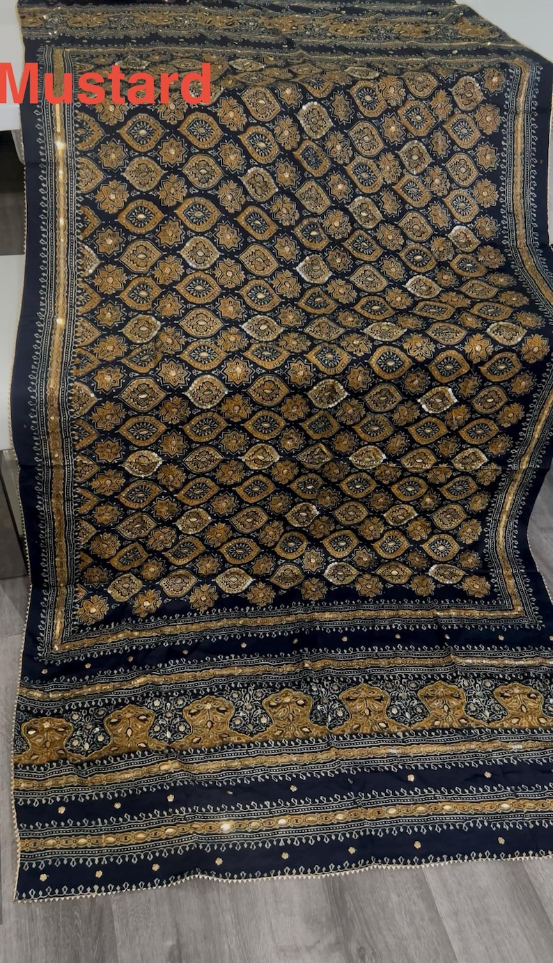 K&ouml;nnte beinhalten: Ein marineblaues Textil mit einem komplizierten Muster aus goldenen und wei&szlig;en floralen und geometrischen Formen. Das Textil hat einen dekorativen Rand und das Wort "Mustard" ist oben links zu sehen.
