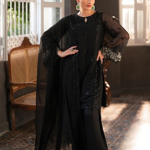May include: A black ensemble featuring a long-sleeved top with intricate embellishments and wide-leg pants. The outfit includes a sheer shawl and is accessorized with green earrings and a ring. A red flower adorns the hair.
